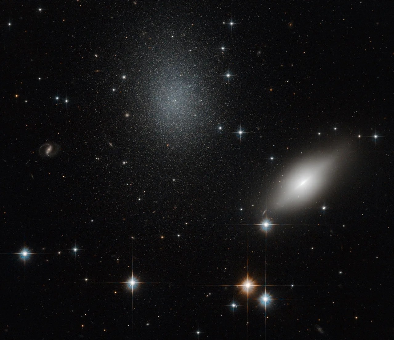 Background stars burn from behind an almond-shaped blurry white galaxy and a diffuse speckled glob of stars