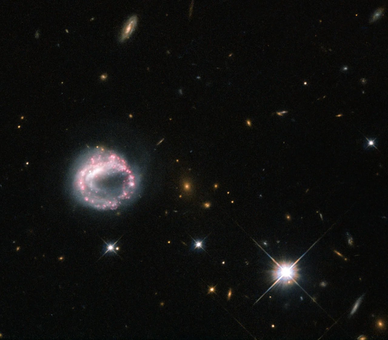 A white circular structure ringed with pink clusters of old stars, a bright star to the right and various distant galaxies