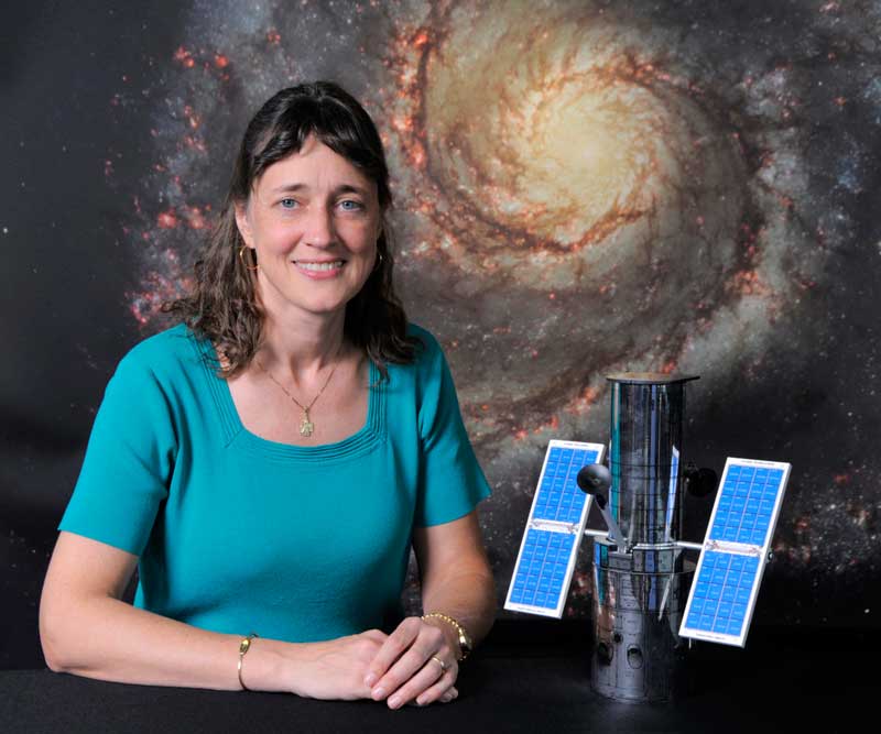 portrait of a woman smiling in front of a Hubble galaxy image. a hubble model on a desk next to her elbow