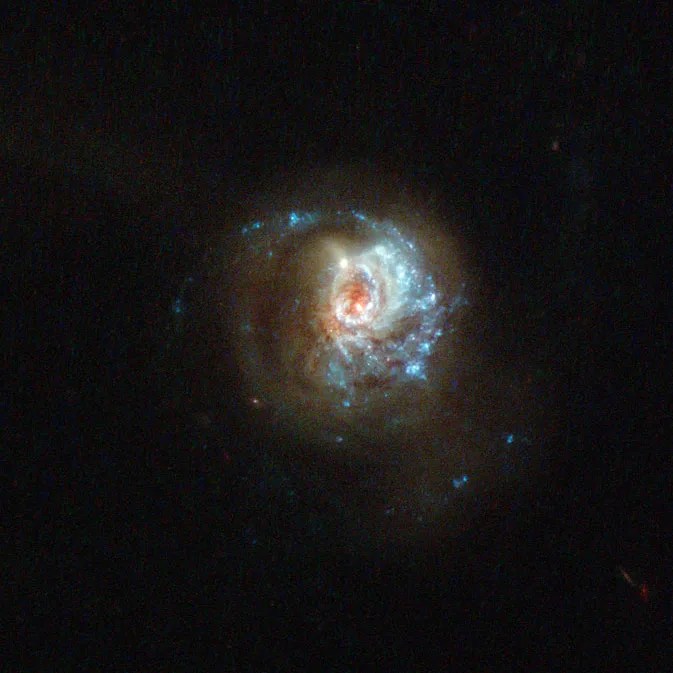 A hazy disk of millions of stars, with an off-center red/white core and brilliant sparkling blue arms stretching out from the bottom, around to the right and trailing up over the top, off into space