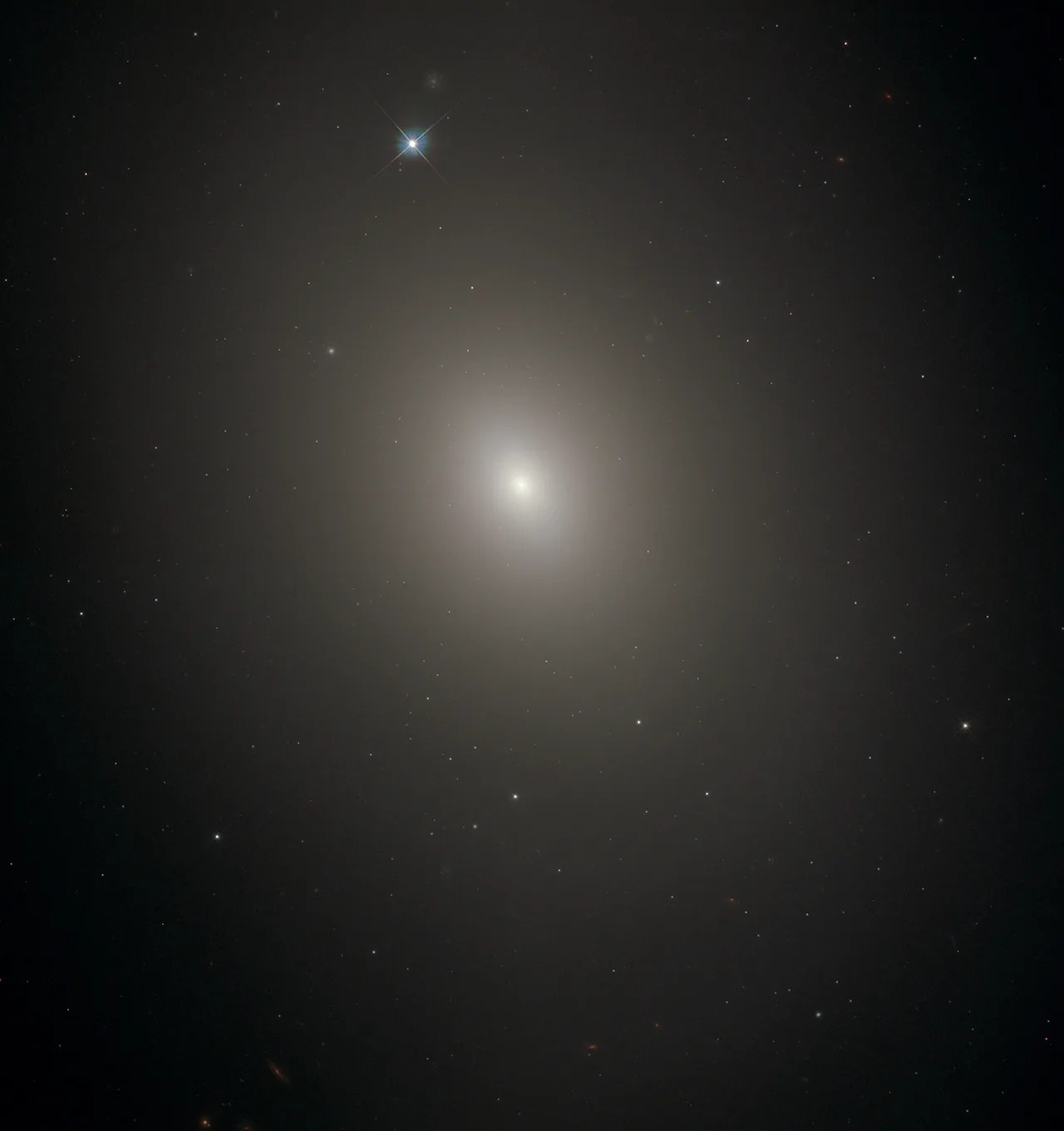 Diffuse oval haze against black background. Stars and distant galaxies shine through it. Just above image center the haze gets brighter and denser toward the bright-white core of the galaxy. Top, just left of center is a bright foreground star.
