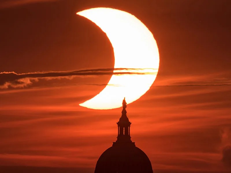 A reddish sky is shown with the bright circle of the Sun, a dark bite taken out of it. The dark circle covering about a third of the Sun's left side is the Moon, during a solar eclipse. In the foreground is the silhouette of the U.S. Capitol.