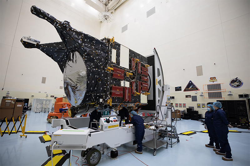 Inside an enormous white-walled clean room, a delivery-truck sized spacecraft sits perched atop a while trailer, and three technicians are working underneath it, while two other workers look on at right, all of them clad in blue coveralls, hair nets, and face masks.