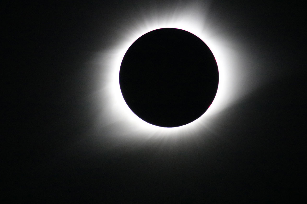 An image of a total solar eclipse during totality, showing a dark circle against a pitch black sky with a white glow surrounding it.