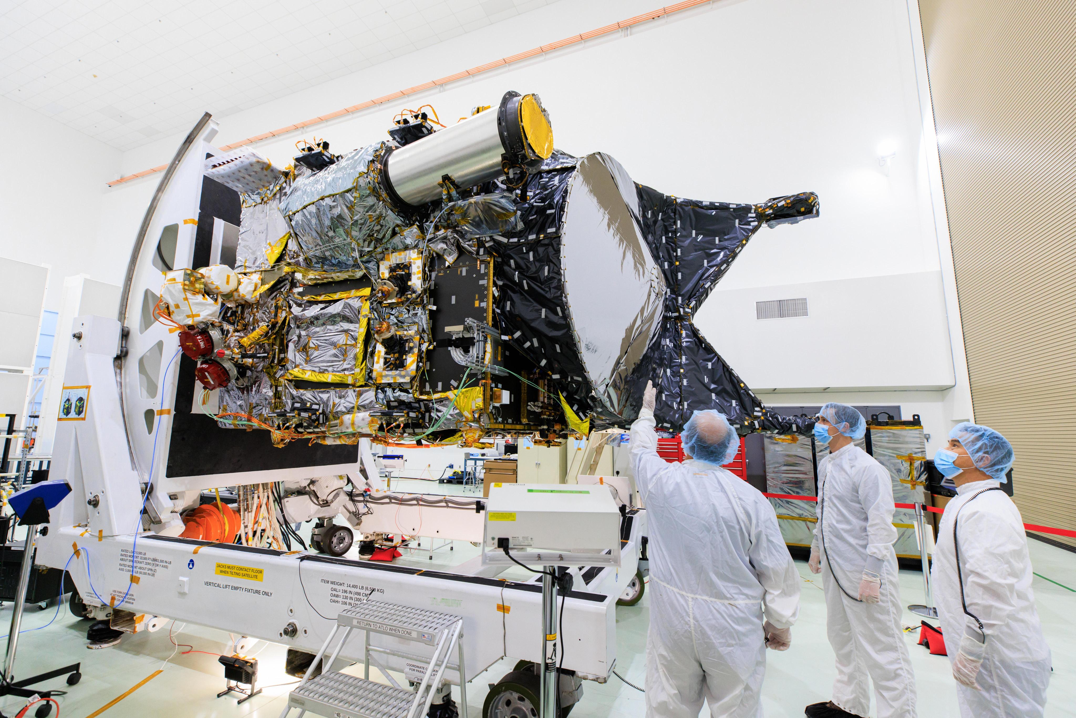 Three workers in white protective gear, hair covers and face mask stand near the Psyche spacecraft as it sits on a stand at Astrotech Space Operations facility.
