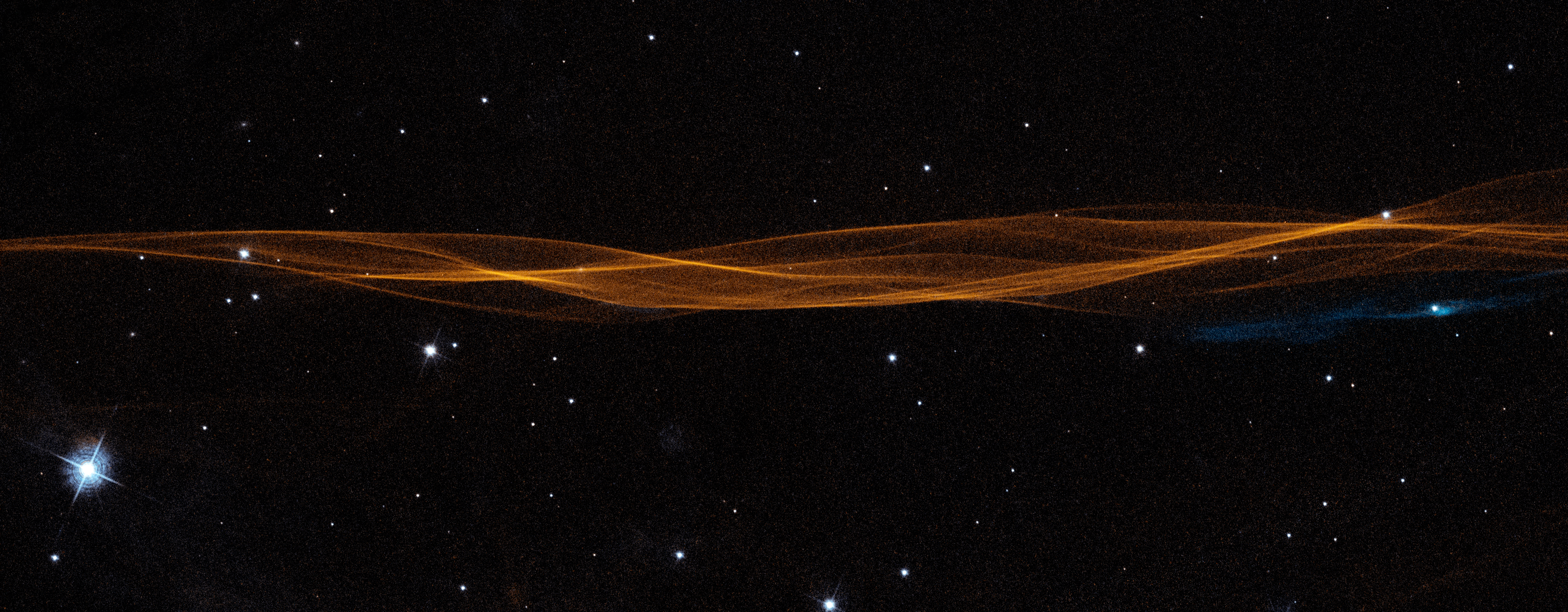 A long, thin, twisted ribbon of orange gas and dust stretches from left to right across the image. Bright-white stars dot the black background. One bright, blue-white star at bottom left. A small swath of blue gas stretches below the orange ribbon on the right side.