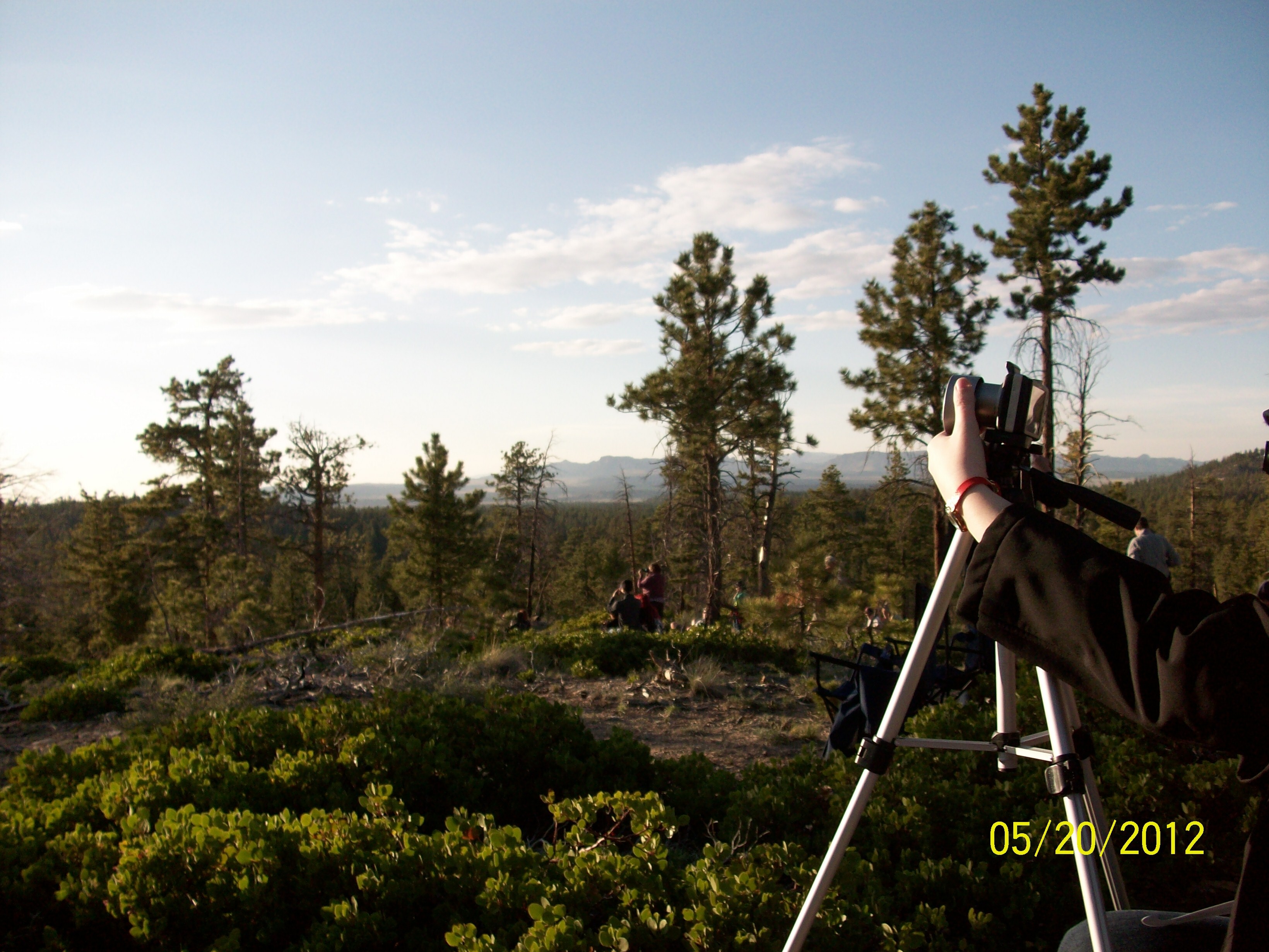 Trees line the landscape. On the right, a camera sits on a tripod. Two hands hold either side of the camera.