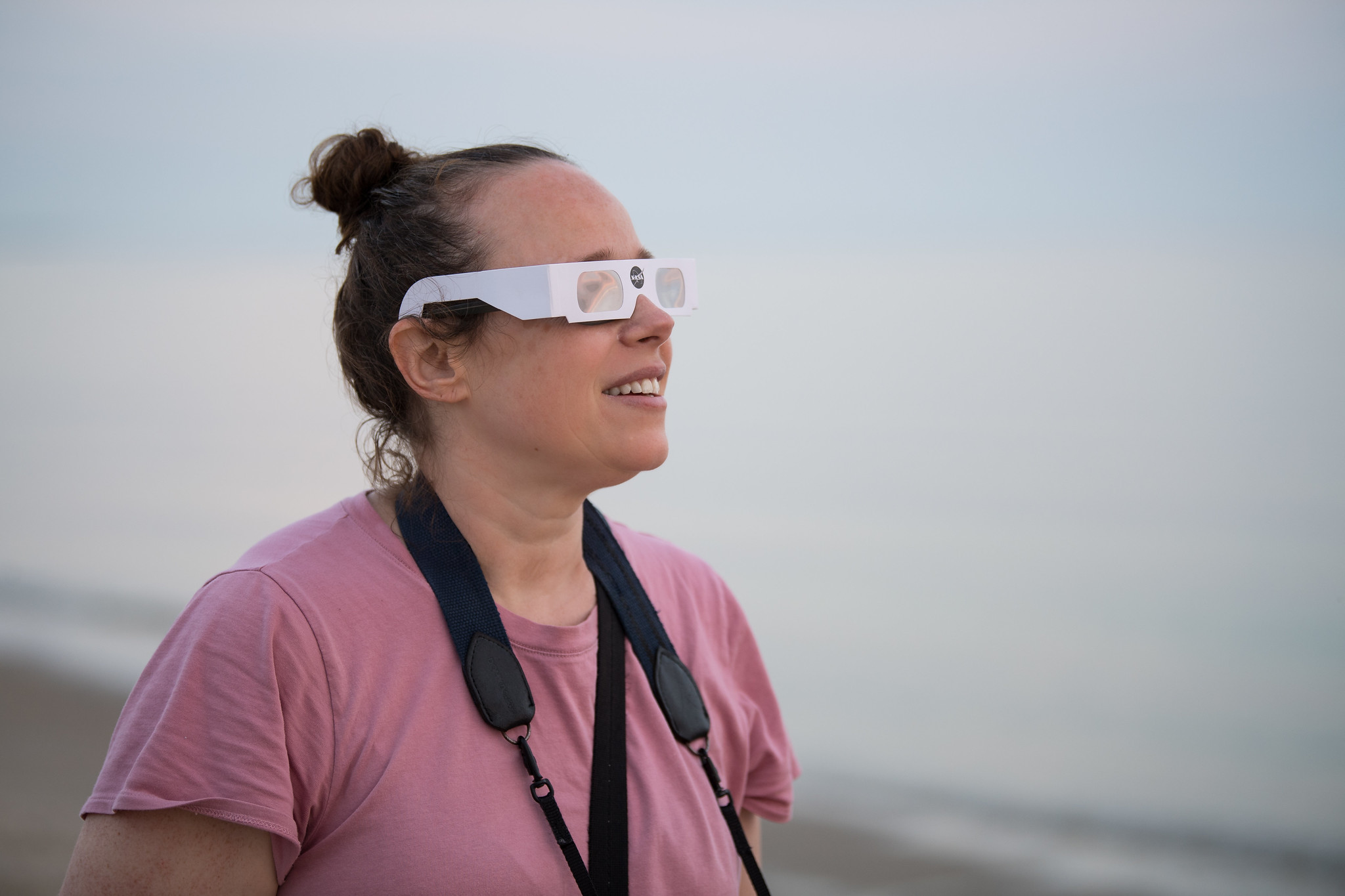 A woman, seen from the torso up, stands on a beach. She is wearing white solar viewing glasses and has a camera strap around her neck.