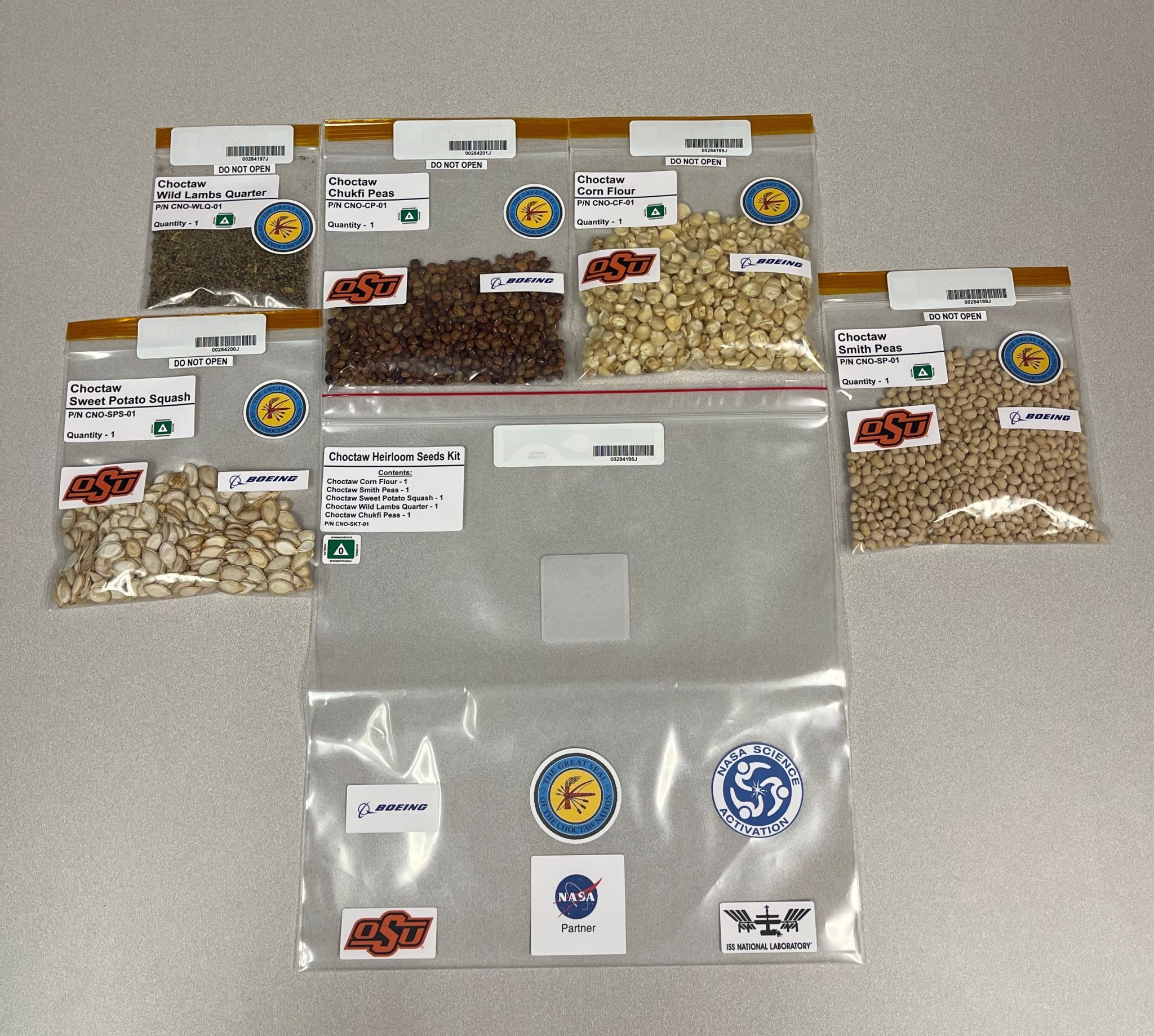 Five clear bags of Choctaw heirloom seeds feature Oklahoma State University and Boeing logo stickers and contain, from left to right, the following kinds of seeds: Sweet Potato Squash, Wild Lambs Quarter, Chukfi Peas, Corn Flour, and Smith Peas. The seeds vary in size, shape, color, and texture, from small, smooth, round, and tan in color, to tiny, rough, teardrop-shaped, and black in color. Below these five bags is a clear bag that will hold them all. This larger bag features the NASA Partner, NASA Science Activation, ISS National Laboratory, The Great Seal of the Choctaw Nation, Boeing, and Oklahoma State University logo stickers.