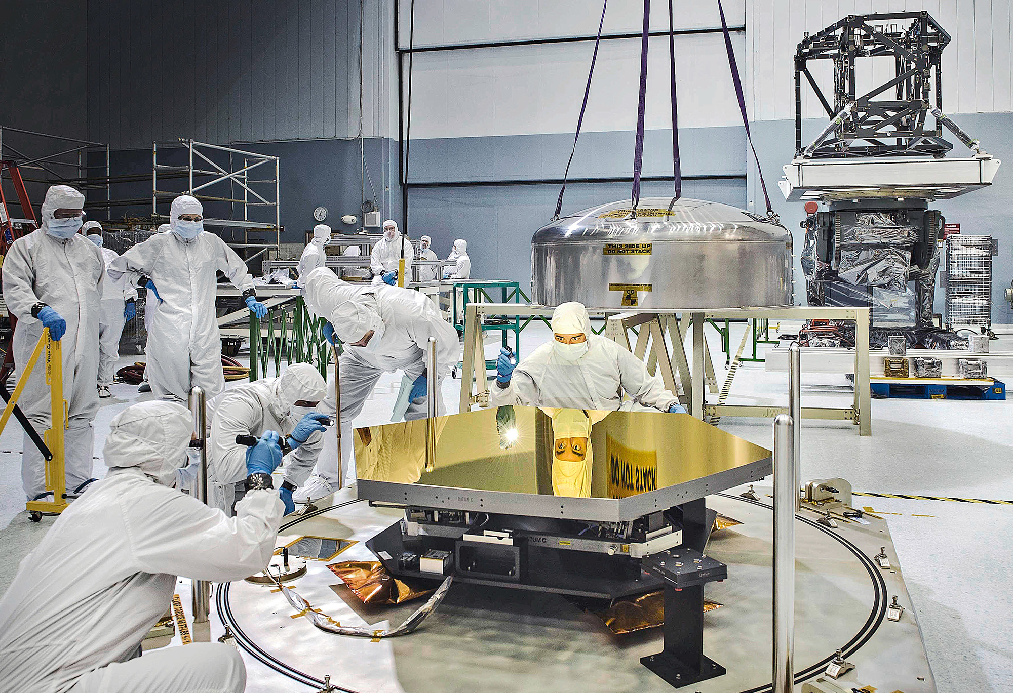 A diverse group of people in white cleanroom suits carefully inspect a single golden faced, hexagonal mirror from the James Webb Space Telescope. Many faces all stare intently, some using flashlights, to examine the mirror surface. Reflected on the surface is the face and intense eye contact from one engineer, and the bright reflection of his flashlight shining directly at the viewer. Behind this engineer, a stainless-steel lens cap used to safely transport the mirror, nearly the size of a human body, rests on scaffolding, still attached by ropes to the crane that lifted it off the mirror moments before. In the background, gray and light blue walls lay behind several other components of Webb scattered around the cleanroom floor. To the right of the frame, a structure made of long overlapping black struts that appear like scaffolding sits on top of a lift table that is meant to safely move the structure up and down.