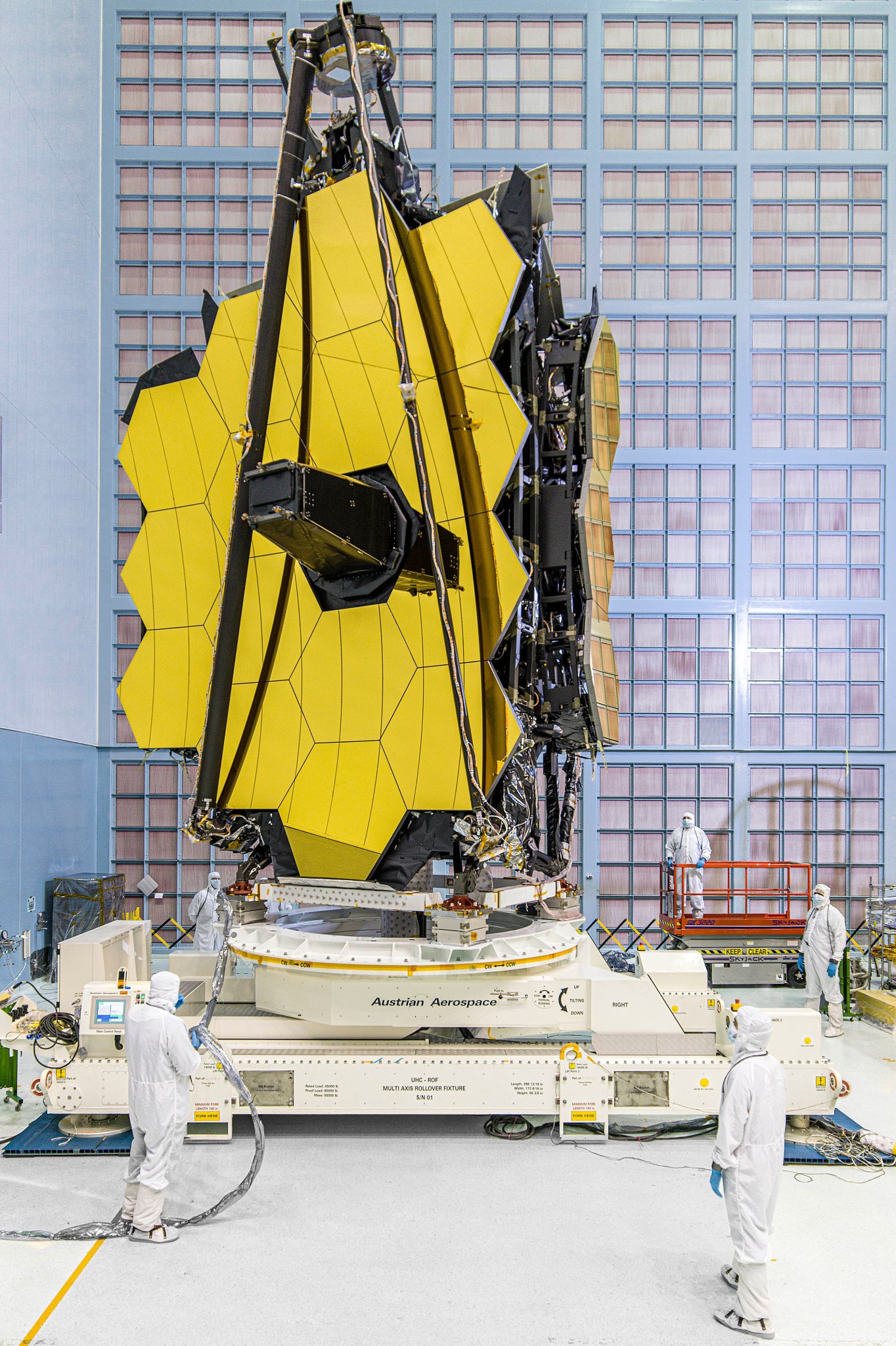 The James Webb Space Telescope mirrors upright on a white stand in the NASA Goddard cleanroom. The 3 mirrors on the right “wing” are folding back like a leaf on a drop-leaf table. The telescope has 18 golden hexagonal segments. The secondary mirror support structure, made of three thin black lines, is folded and the secondary mirror sits atop this folded tripod. There are 5 people in white cleanroom suits supervising the folding maneuver. One of them is on a red lift at the back right. There is a wall of HEPA filters behind the telescope.