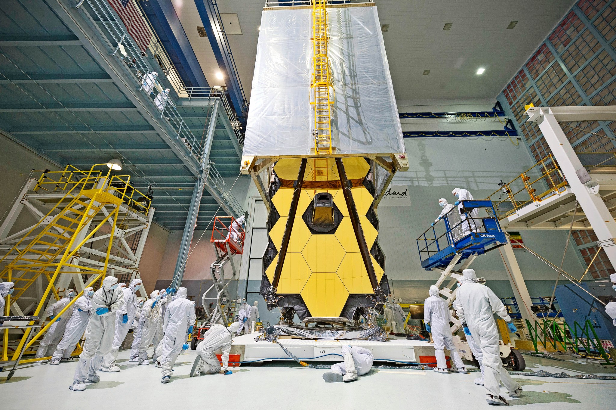 The Webb telescope mirrors are shown in the NASA Goddard cleanroom, with a clean tent, which looks like a giant rectangle made of clear plastic tarp, half over the mirrors. A crane is lowering it down. The mirrors are gold hexagons. The three on each side wing are folded back like a drop leaf table. The telescope is being prepped for vibration testing. For testing, it has to be removed from the cleanroom, so the tent will protect it while it is being tested. There are multiple people in white cleanroom suits that cover them from head to toe and protect the cleanroom from hair and skin cells and contaminants on clothes. Two of the people are on a blue lift to the right of the telescope, and one on a red and silver left to the left, inspecting the work. There is a yellow ladder at far left. There are various white support structures. The wall at far right is covered in HEPA filters. The very large cleanroom door is directly behind the telescope.