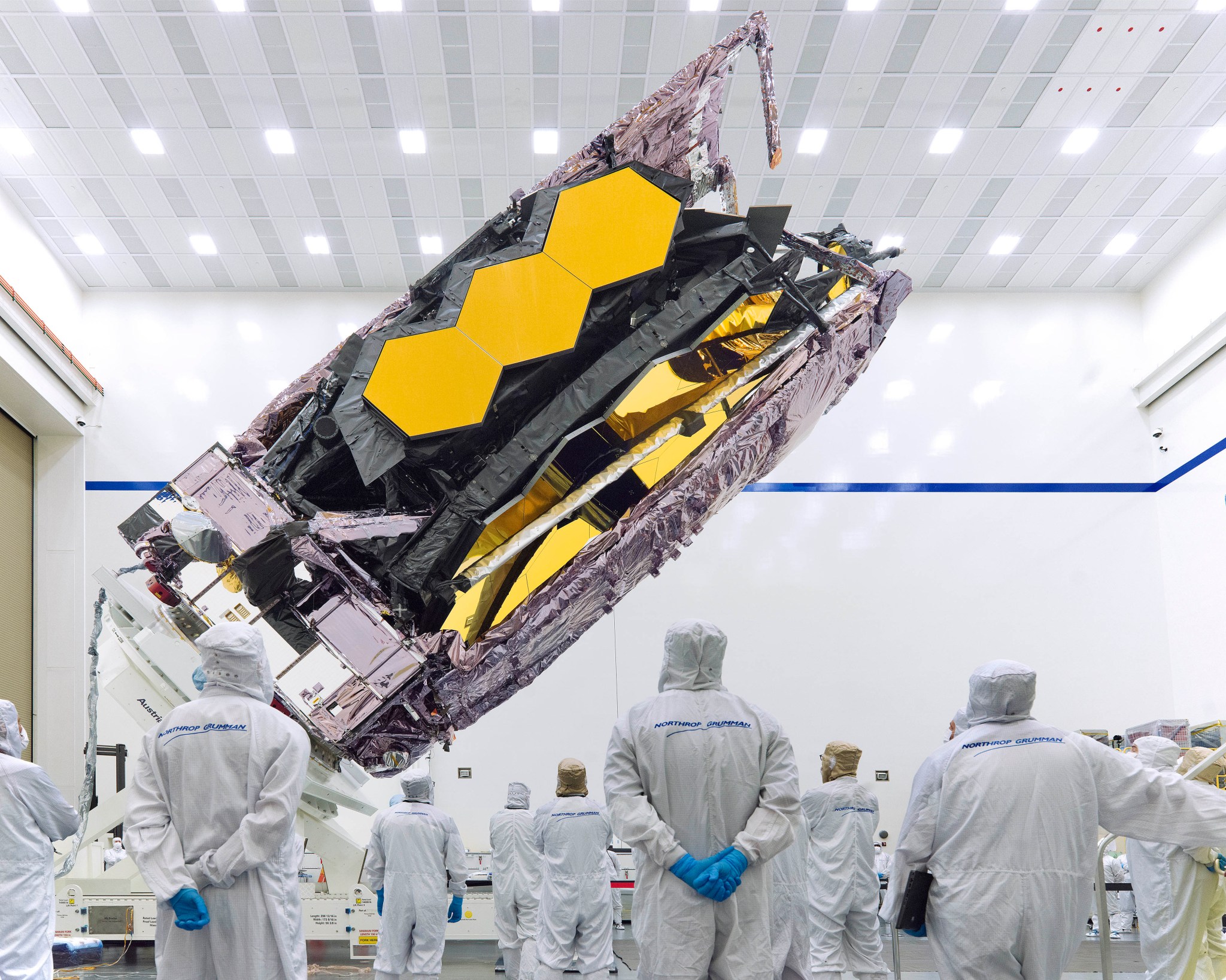 Several cleanroom technicians stand and watch as the Webb telescope, in its folded configuration, is being lifted and horizontally tilted towards the right side. When folded, Webb’s purple, metallic pallets surround its hexagonal, gold-coated mirror segments, making a long rectangle shape. The technicians wear blue gloves and white contamination-control suits that have the blue Northrop Grumman logo on the back. This image was taken in the Northrop Grumman cleanroom, which features a blue line running across its bright white walls.