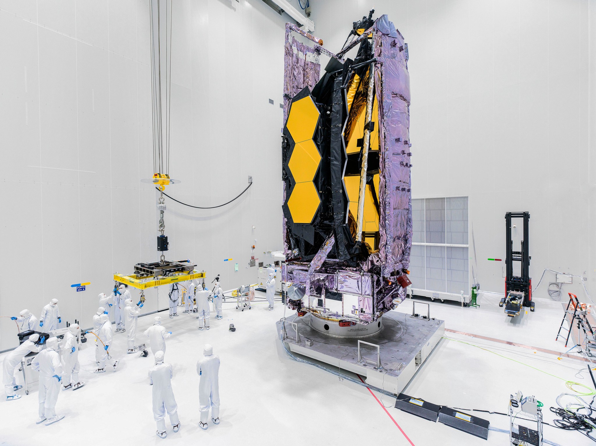 Image taken in a cleanroom at Europe’s Spaceport in Kourou, French Guiana. Dozens of cleanroom technicians, wearing white contamination-control suits and blue gloves, stand towards the left side of this image. On the right side is the Webb telescope in its folded configuration. Its purple sunshield pallets enclose its hexagonal, gold-colored mirror segments. Other hardware, wires, and equipment are located around the cleanroom, which features bright white walls.