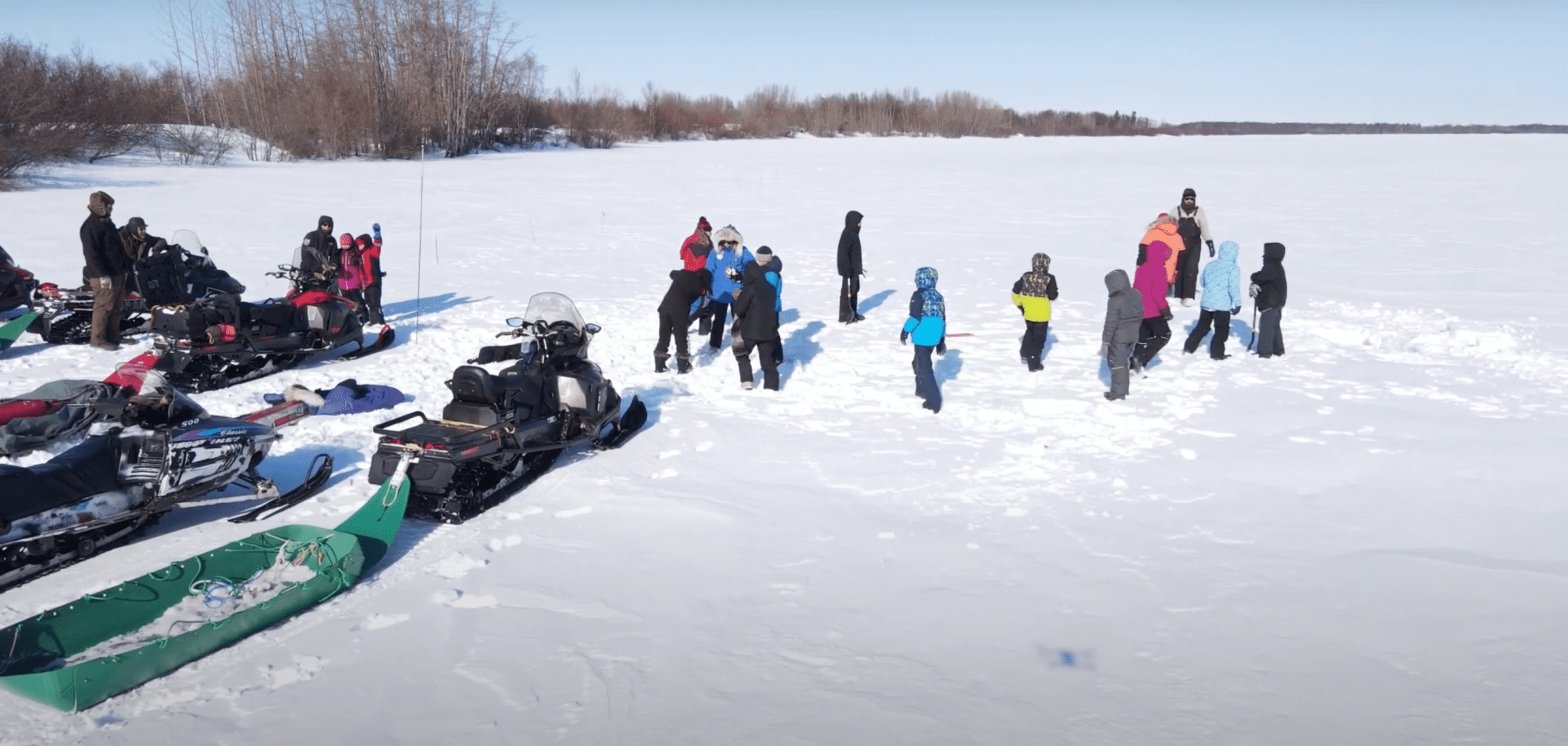 Photograph of about 20 people in colorful jackets gathered on lake ice. Snowmobiles, backpacks and other equipment are nearby.